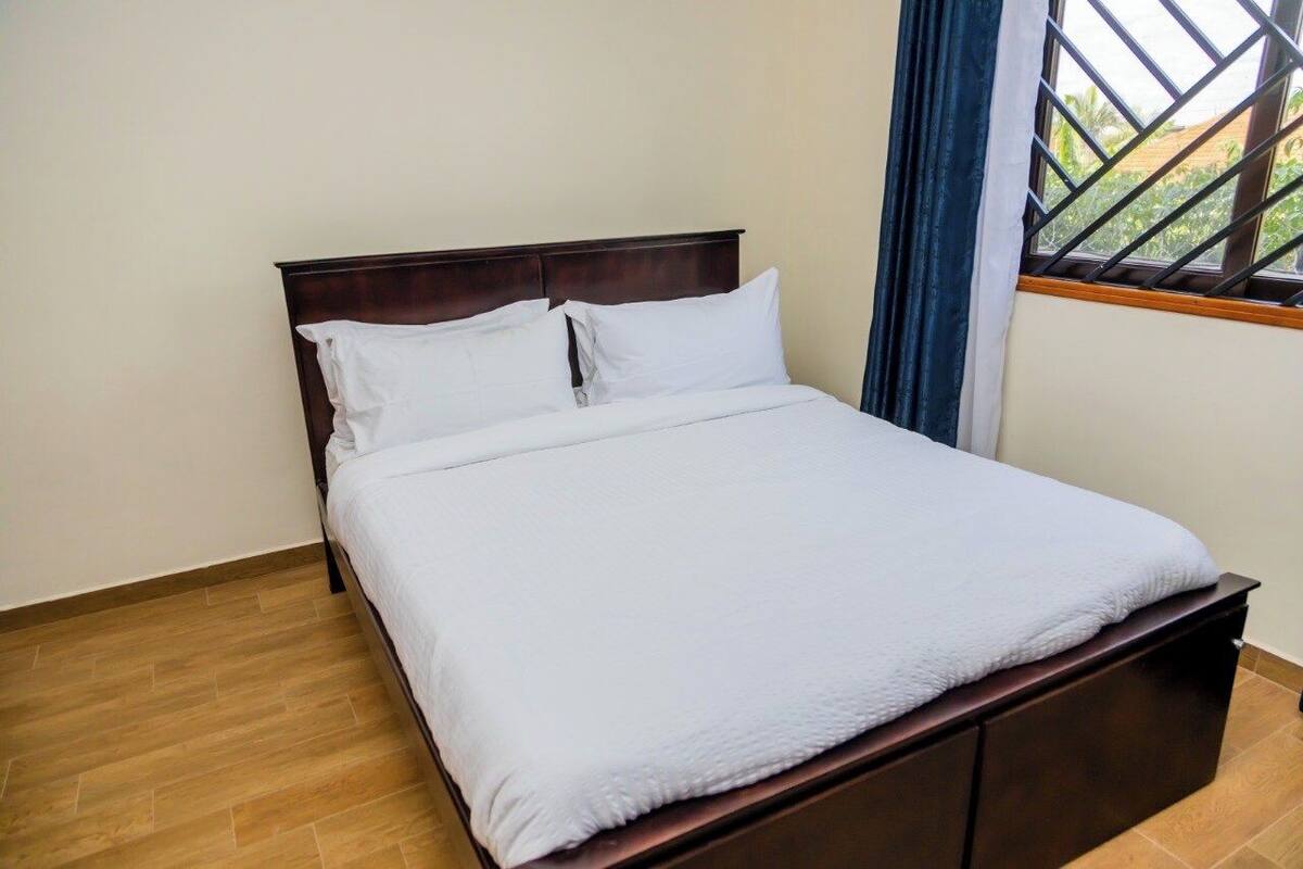 A neatly made bed with white bedding in a room with wooden flooring and a window with a metal grid.