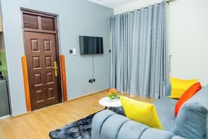 A living room with a brown door, a flat-screen TV mounted on the wall, a grey sofa with yellow and orange cushions, and a small round table with a flower vase. - Cozy Residences Kampala 005 (Kampala)