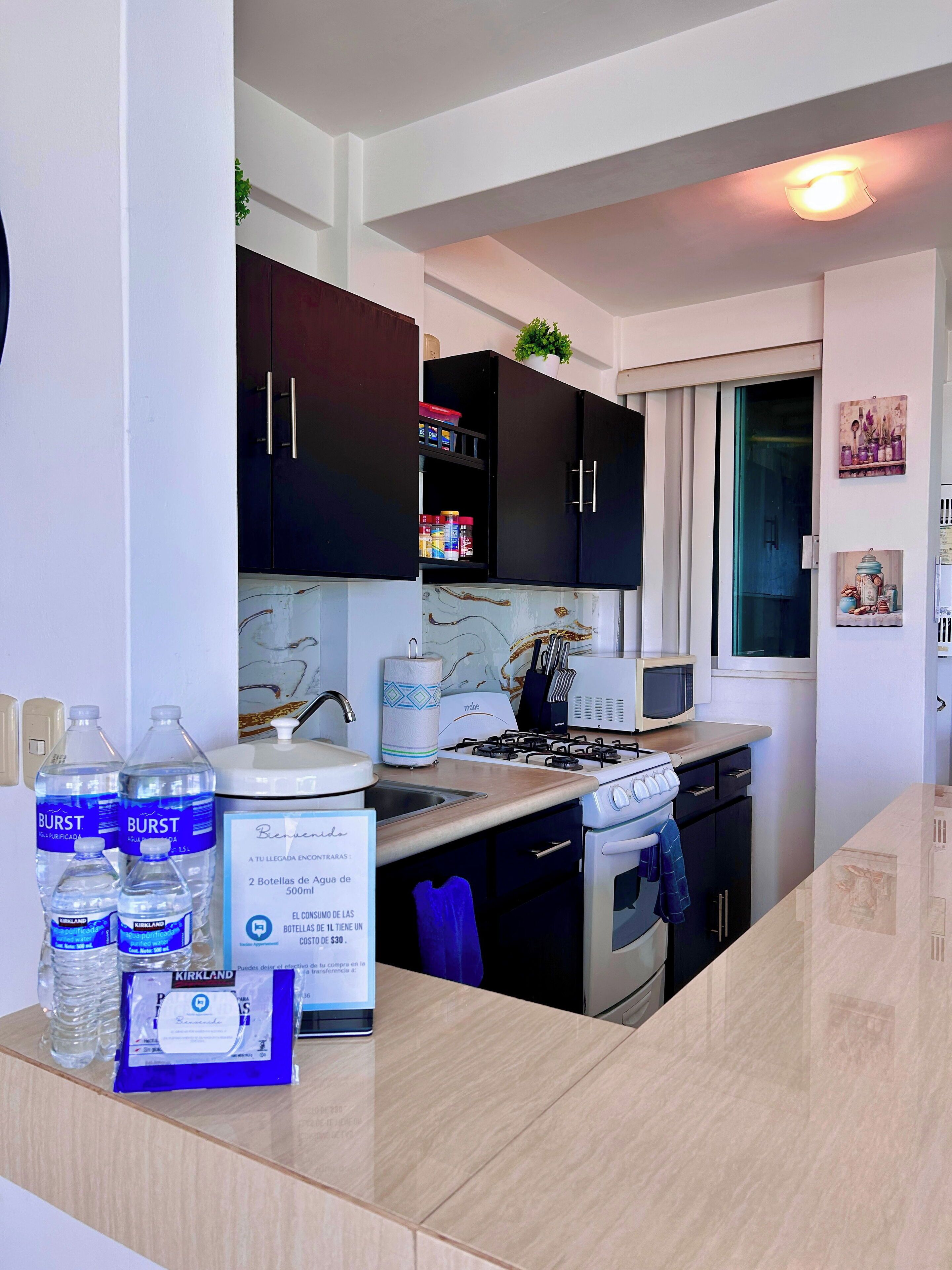 A modern kitchen with dark cabinets, a microwave, a stove, and a sink. There are water bottles and a Kirkland brand package on the counter.