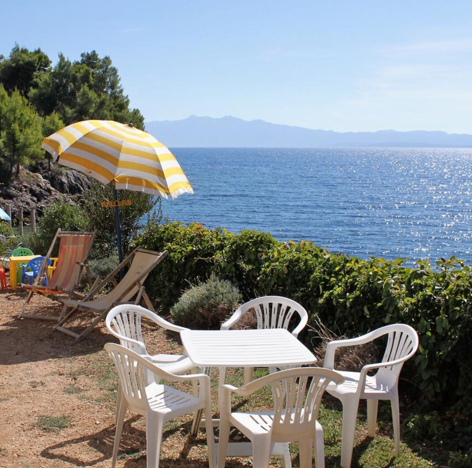 Une terrasse avec des chaises et une table en plastique blanc, d’où l’on peut admirer un plan d’eau avec des montagnes à l’horizon.