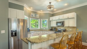 A kitchen with white cabinets, granite countertops, a stainless steel refrigerator, a microwave, and a water dispenser. There are wooden bar stools and a chandelier.