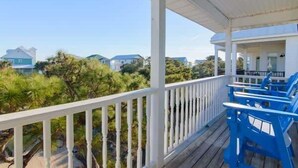 A covered porch with white railings and blue chairs, overlooking a residential area with houses and greenery.