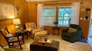 Una sala de estar acogedora con paredes de madera, un sillón verde, un sillón con estampado floral, una mesa de centro de madera y un sofá verde.