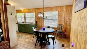 Un área de comedor con una mesa redonda, cuatro sillas y una vela azul. Hay una zona de cocina con un fregadero y armarios verdes.