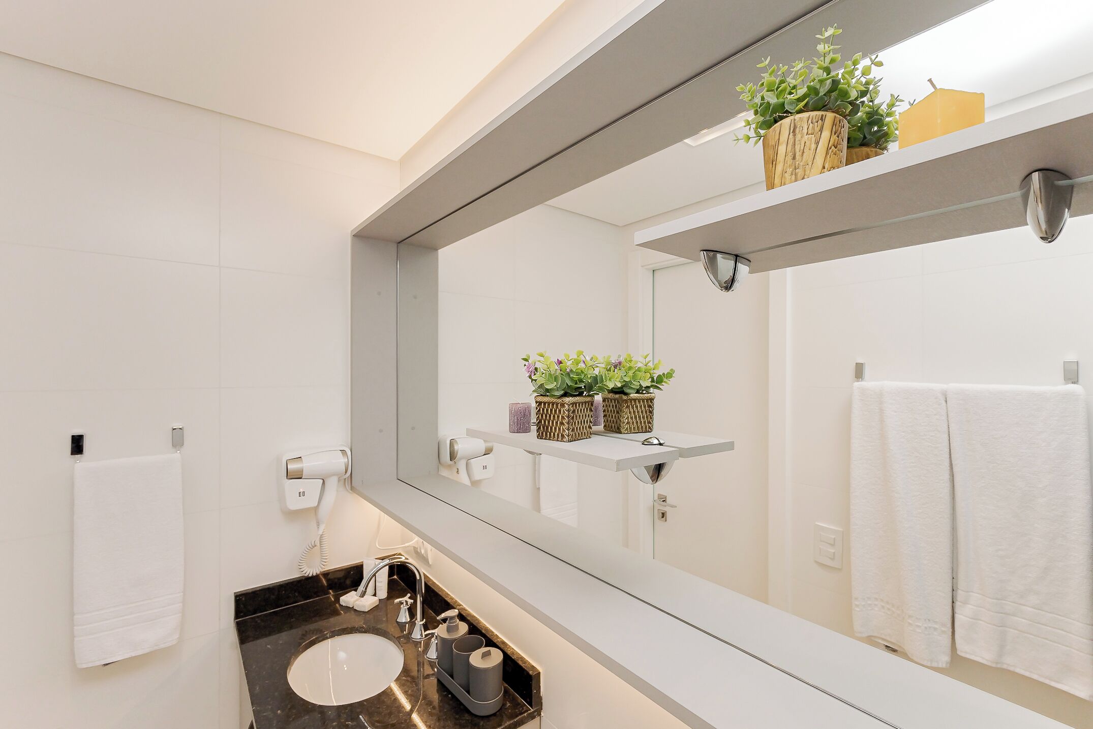 A modern bathroom with a large mirror, white tiles, a black sink, and a shelf with plants.