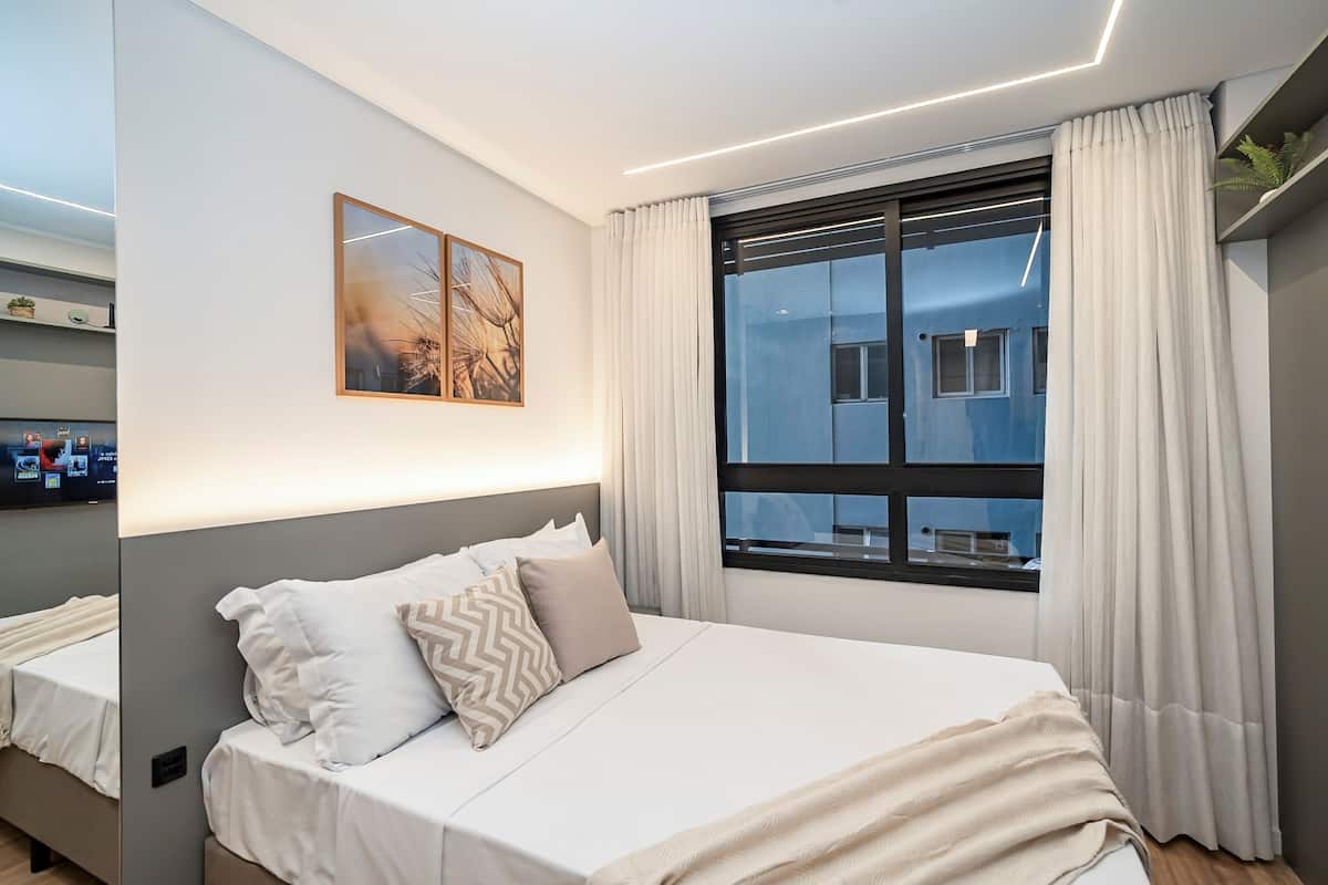 A modern hotel room with a large bed, a window with curtains, a wall-mounted TV, and a framed picture.