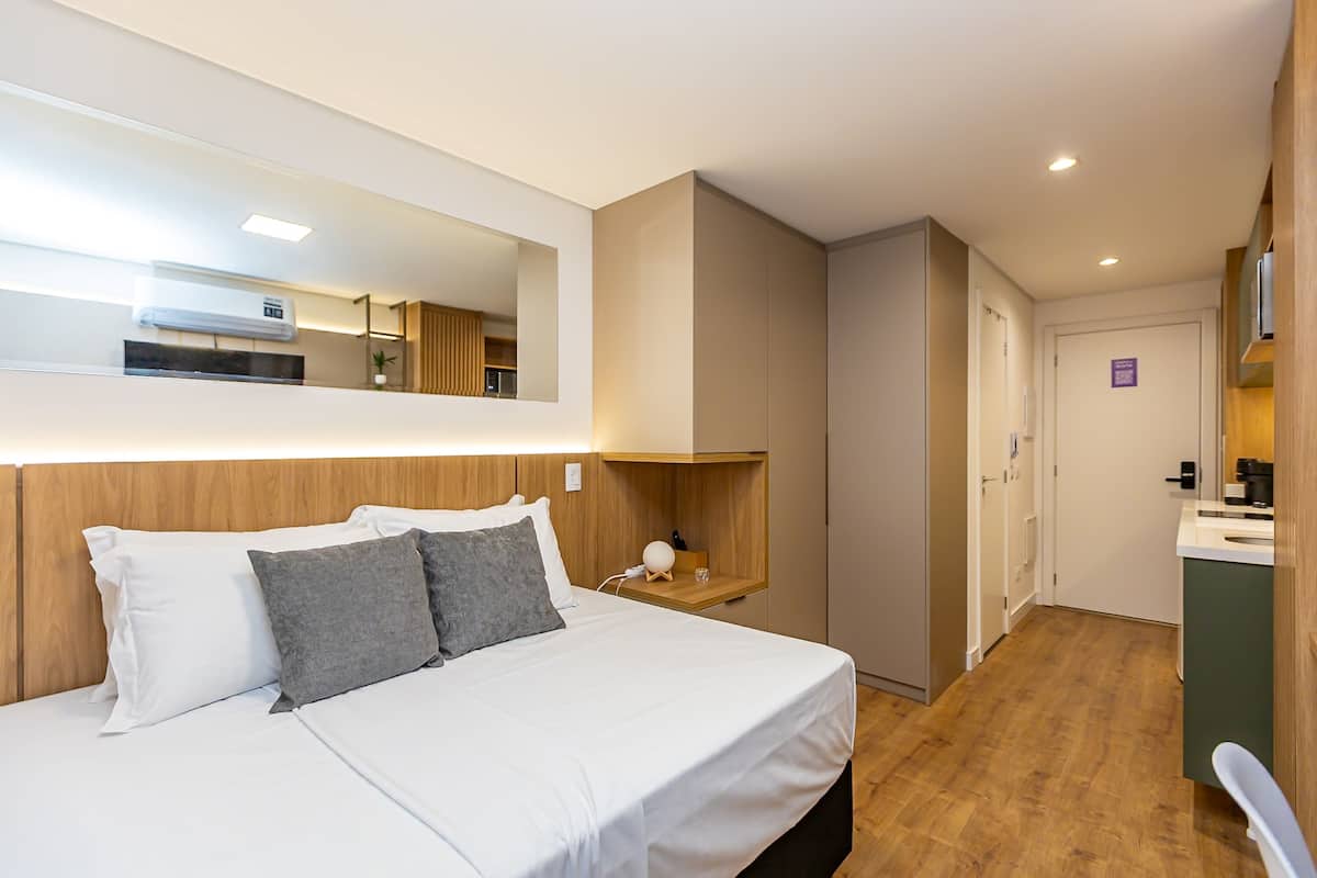 A hotel room with a bed, a bedside table, a mirror, a small shelf, and a view of the kitchen and bathroom.