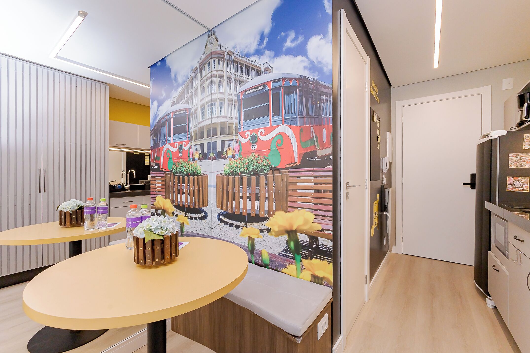 A modern hotel room with a large mural of a tram, two round tables with flower arrangements, and a wooden bench.