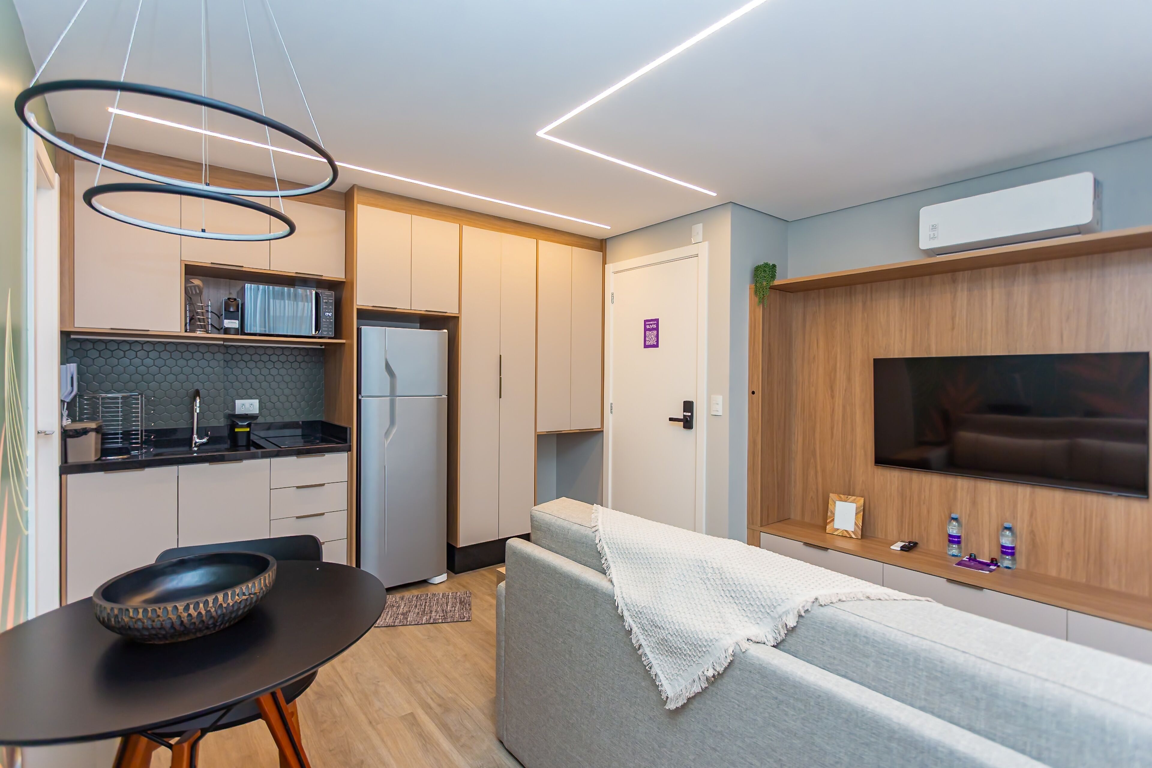 Ett modernt kök med ett svart matbord, en mikrovågsugn och ett kylskåp. Det finns en grå soffa med en vit pläd och en tv på väggen.