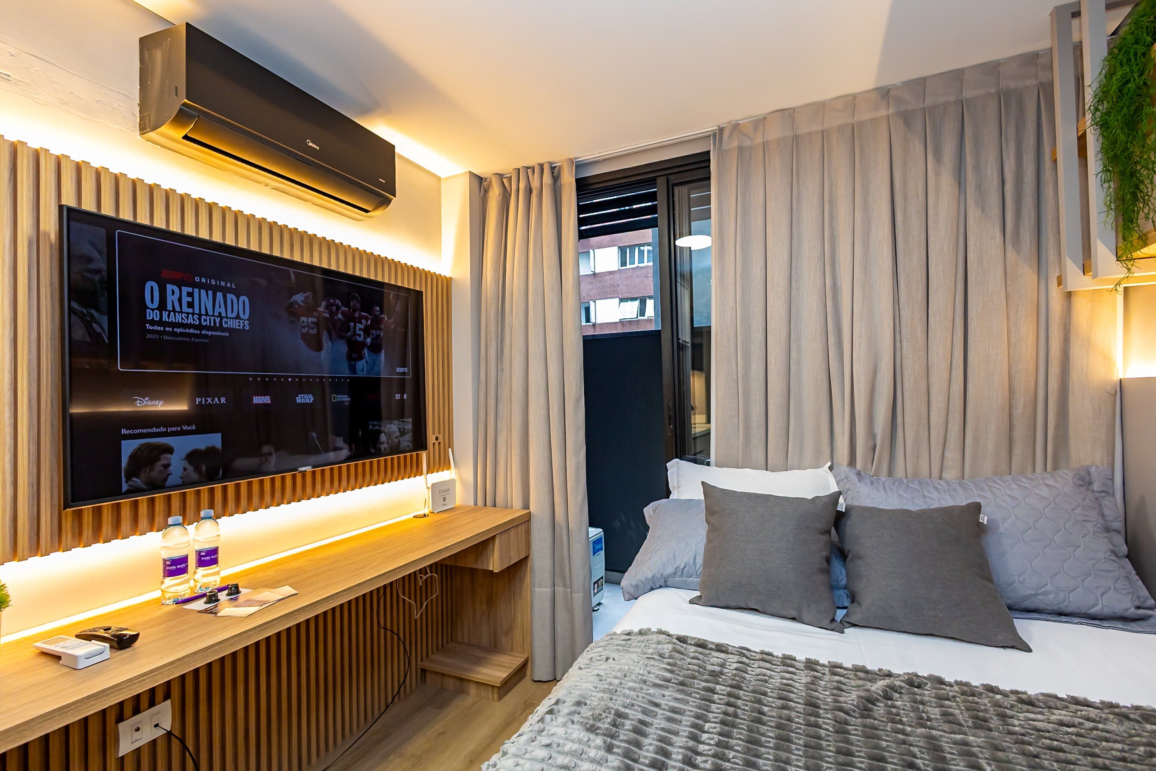 A modern hotel room with a large flat-screen TV mounted on the wall, a wooden desk with a lamp, and a bed with grey and white bedding.