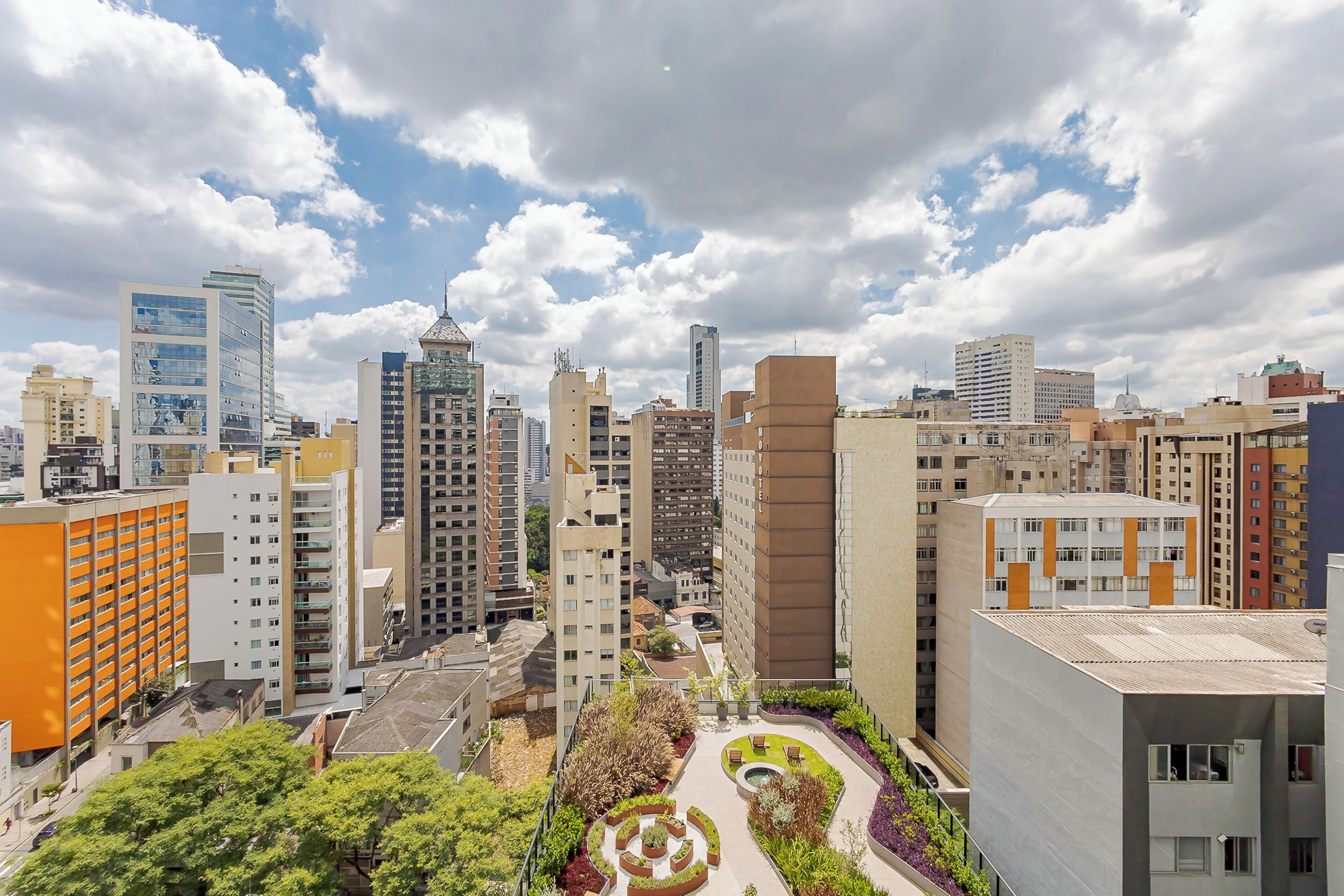 Une vue urbaine avec des immeubles modernes de grande hauteur, une cour centrale aménagée avec des sièges et un ciel dégagé.