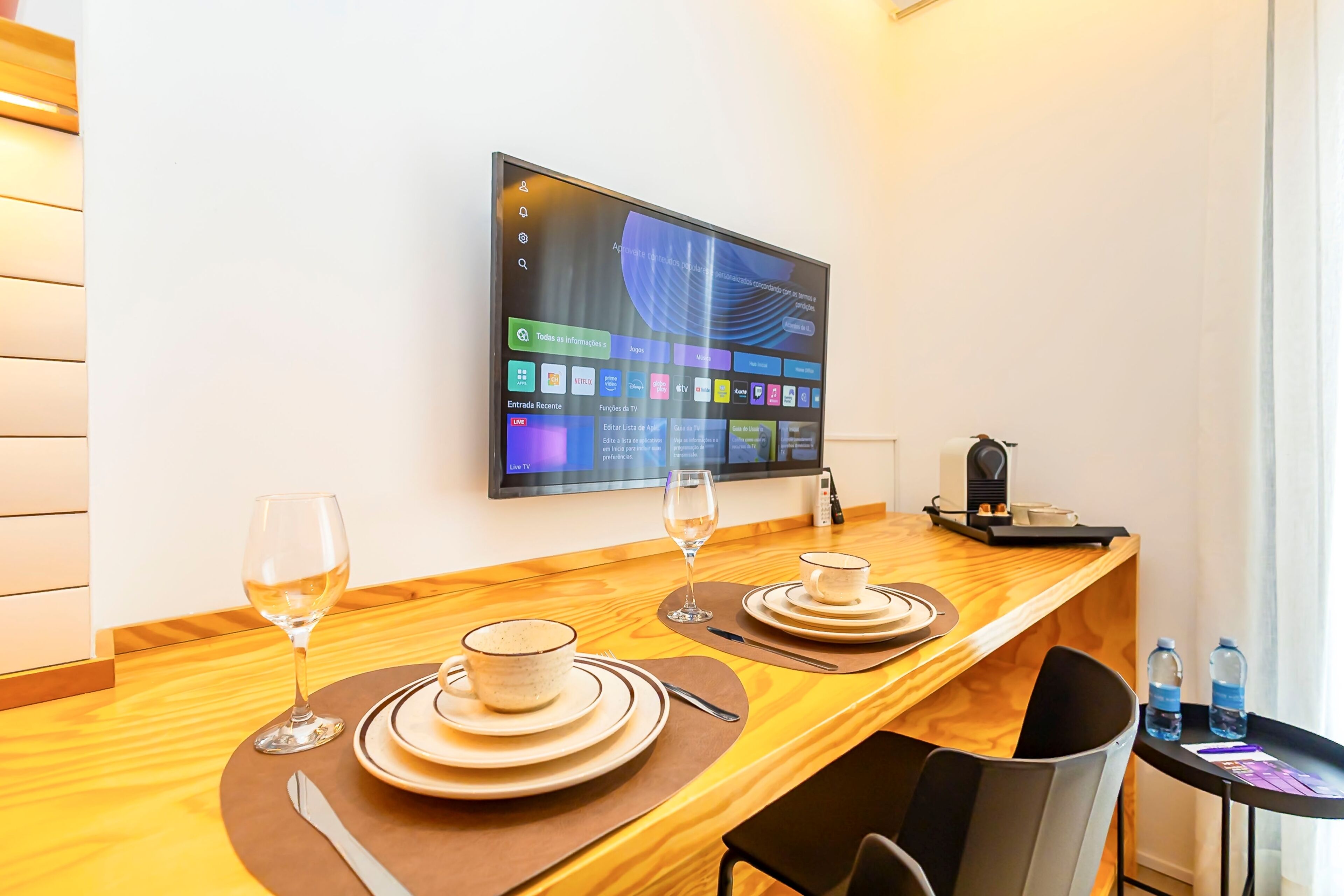 Een hotelkamer met een houten eettafel gedekt voor twee personen, een flatscreen televisie aan de muur en een klein bijzettafeltje met waterflessen.