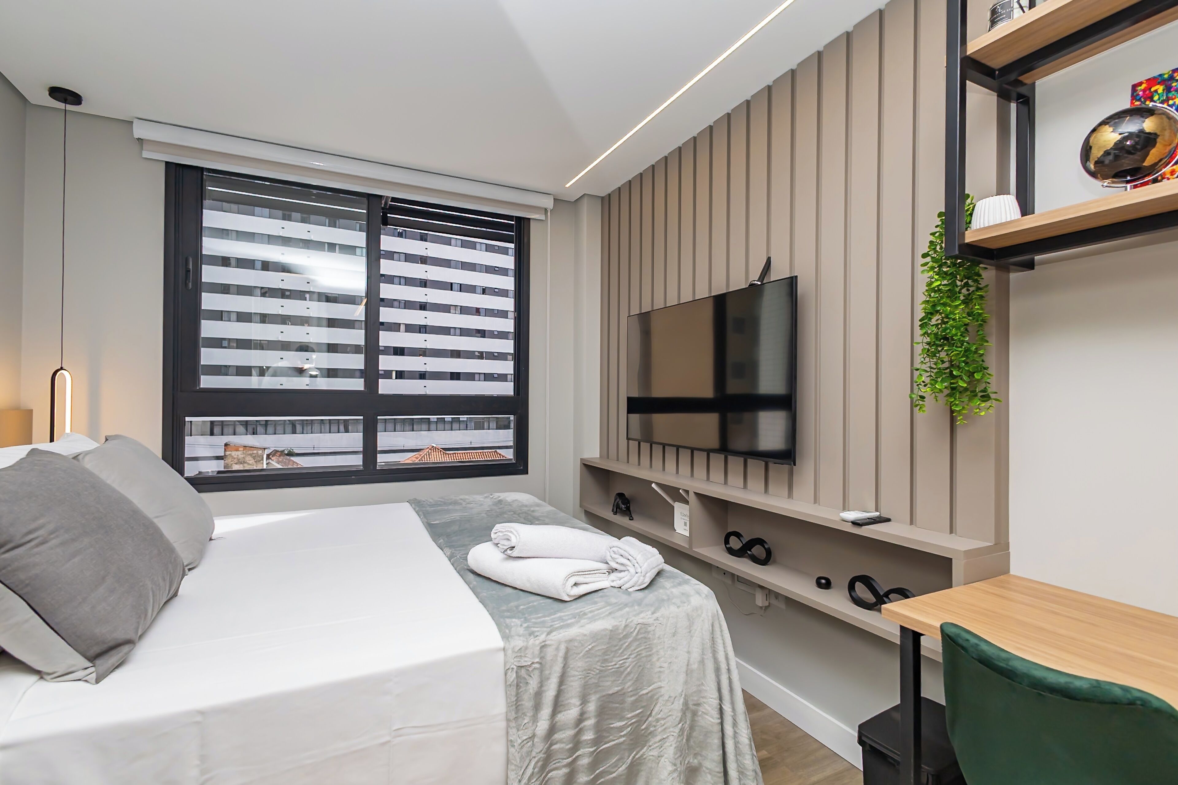 Et moderne hotelværelse med en seng, et fjernsyn monteret på væggen, et lille bord og et vindue med persienner.