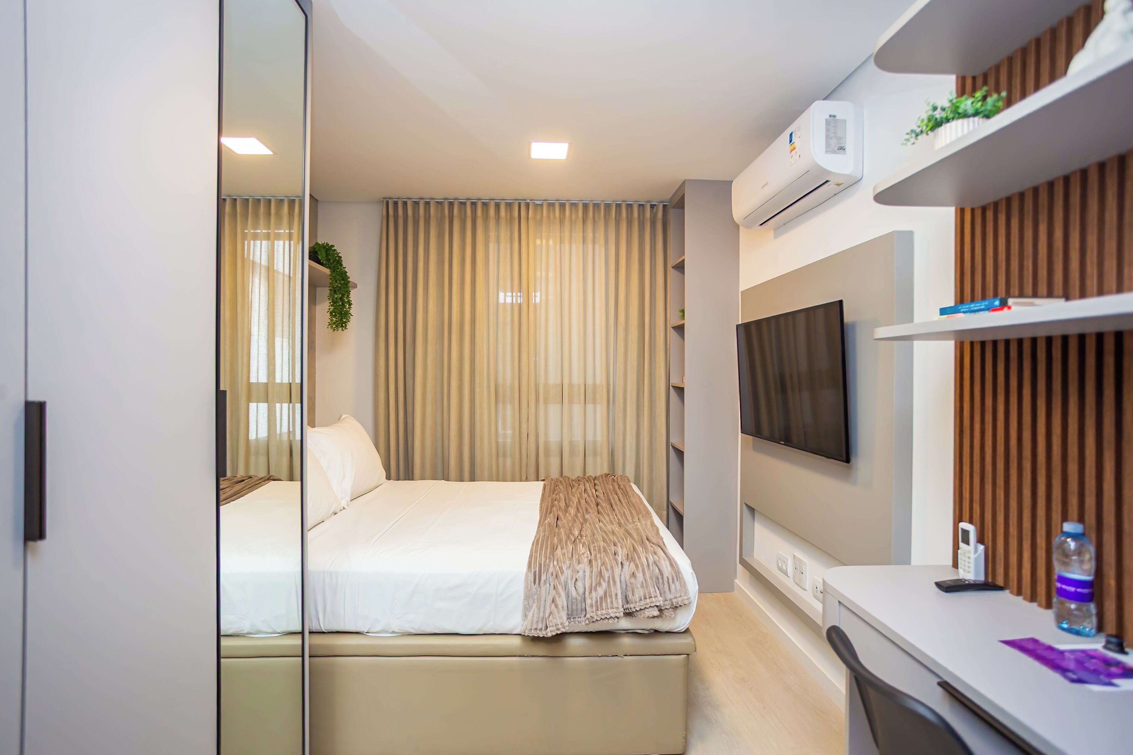 A modern hotel room with a bed, bedside table, TV, and air conditioning unit.