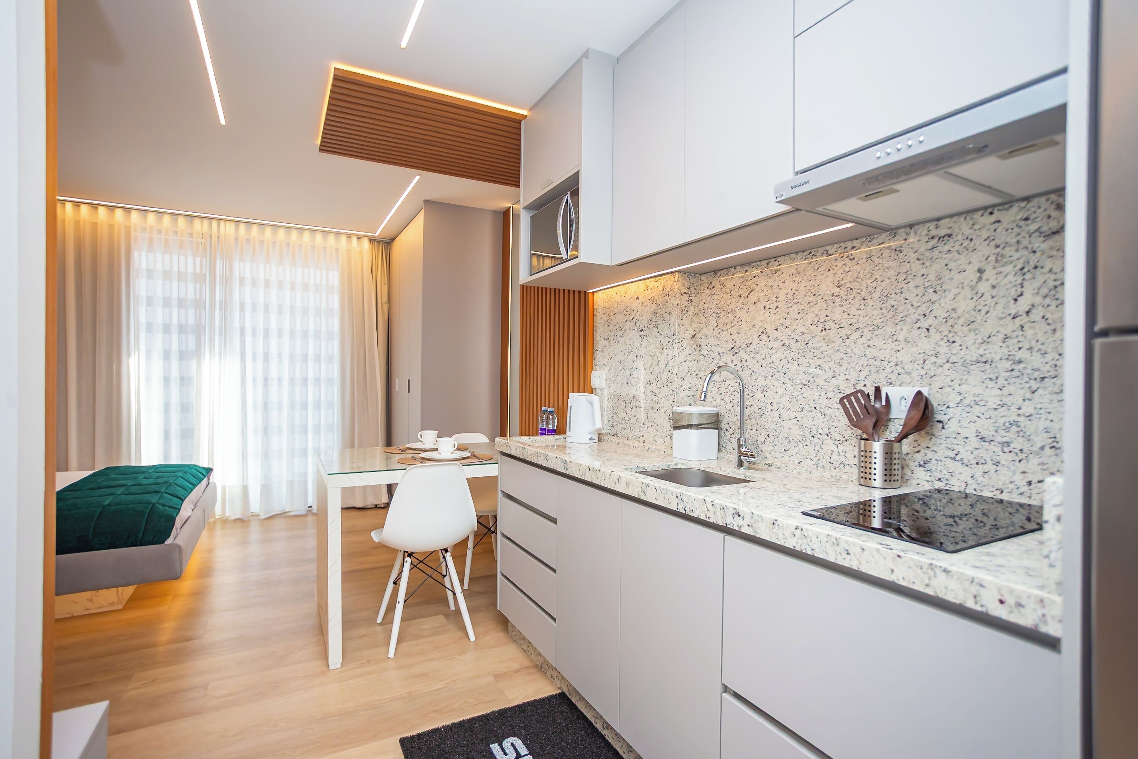 A modern kitchen with a granite countertop, stainless steel sink, and a microwave. There is a dining area with a white table and chairs, and a bed with a green blanket in the adjacent room.
