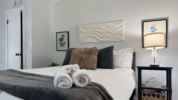 A neatly made bed with a grey blanket, two rolled towels, and a brown pillow. There is a framed picture of flowers on the wall, a lamp on a side table, and a small basket on the table. - Downtown EVV Studio / Chandler C (Evansville)