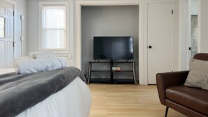 A bedroom with a bed, a TV on a stand, a brown leather sofa, and a door. - Downtown EVV Studio / Chandler C (Evansville)