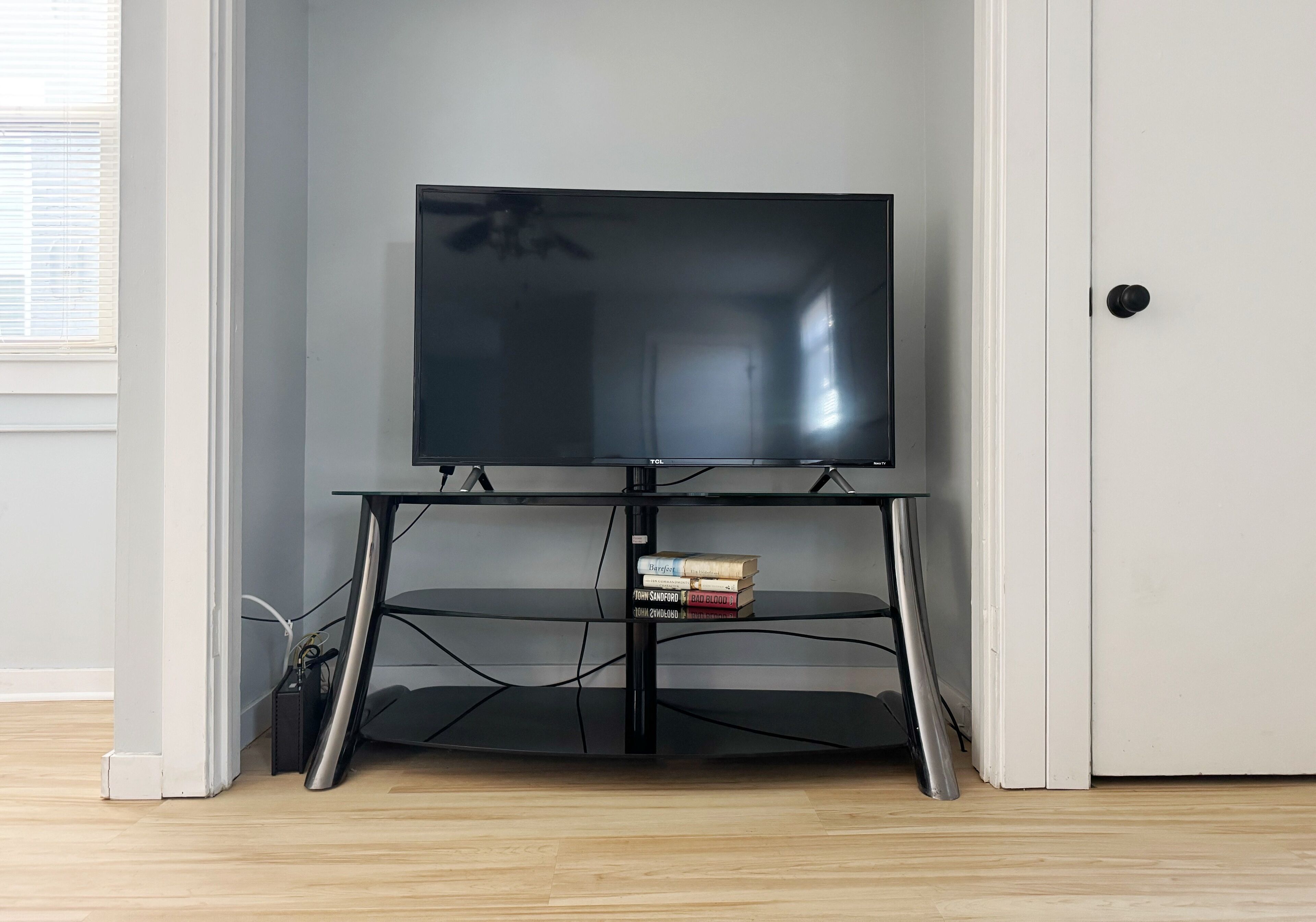 Een flatscreen televisie op een zwarte tv-standaard met een plank eronder, in een kamer met houten vloer en een witte deur.