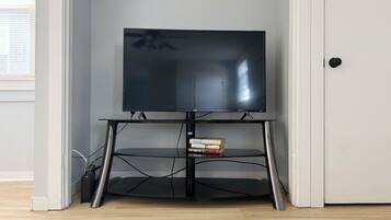 Een flatscreen televisie op een zwarte tv-standaard met een plank eronder, in een kamer met houten vloer en een witte deur.