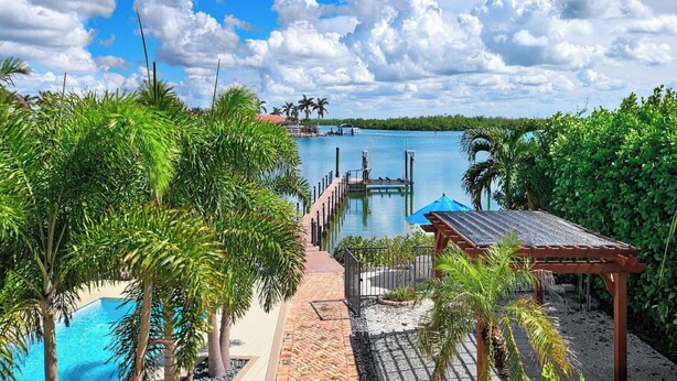 A pool area with a dock extending into a body of water, surrounded by lush greenery and a clear sky.