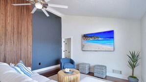 A modern living room with a large bed, a ceiling fan, a wall-mounted TV displaying a beach scene, and a potted plant.