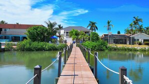 A wooden pier leading to a house with a covered area, surrounded by greenery and palm trees.