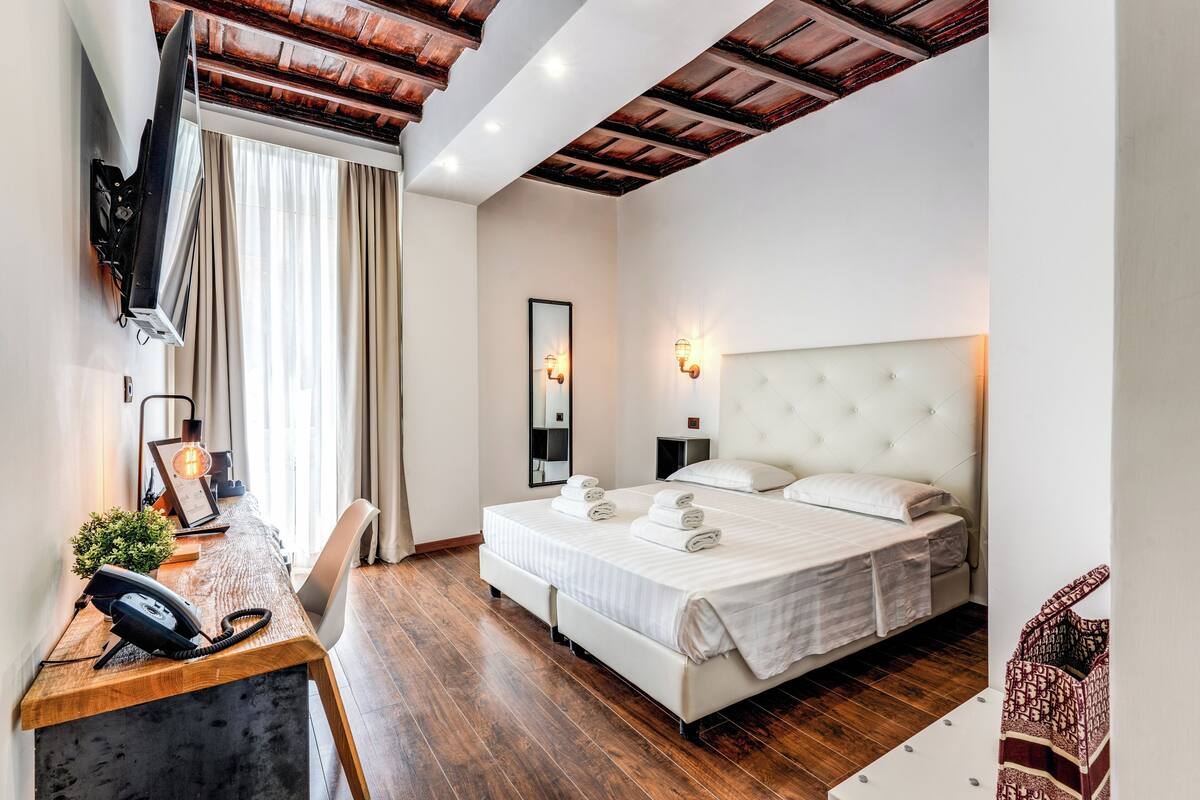 Ein Hotelzimmer mit einem Holztisch, einem Bett mit weißer Bettwäsche, einem an der Wand befestigten Fernseher und einem großen Spiegel.