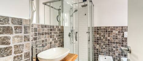 Een moderne badkamer met een backsplash van stenen mozaïek, een douche met glazen wanden, een wit wastafelmeubel en een bidet.