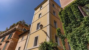 Ein mehrstöckiges Gebäude mit gelber Fassade und grünen Fensterläden, mit einer Straßenlaterne und üppigem Grün.