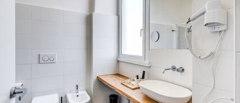 A modern bathroom with a wooden vanity, a white sink, a mirror, a toilet, and a hairdryer mounted on the wall.