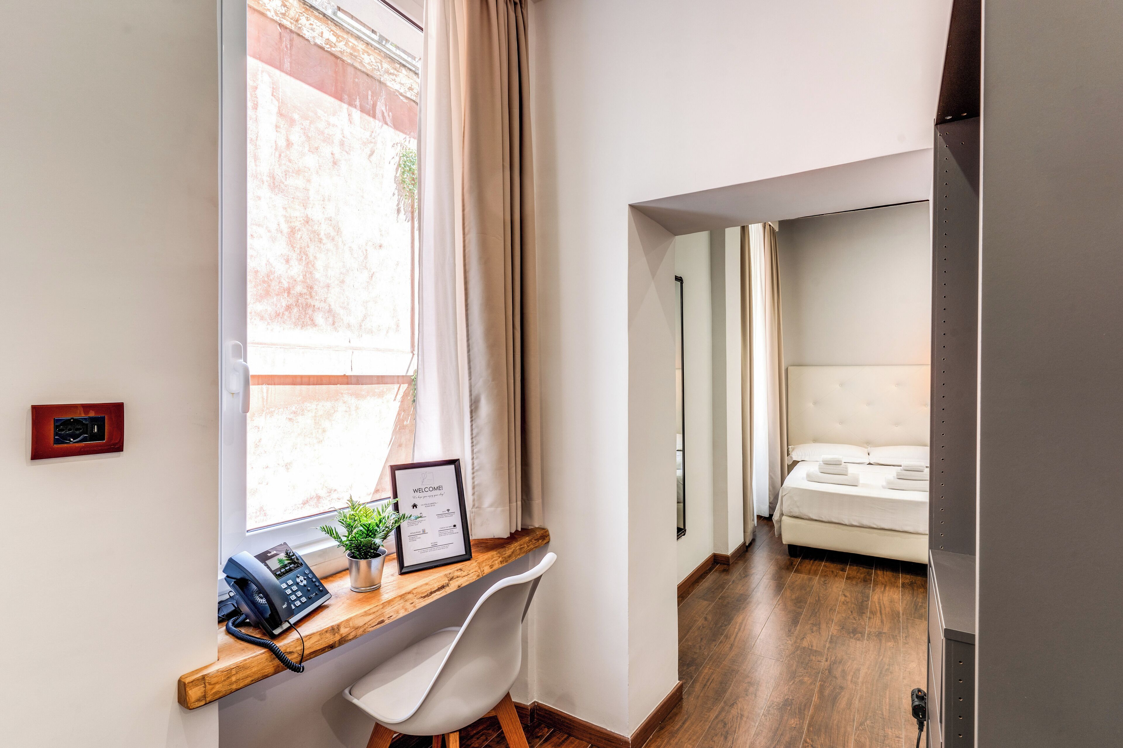 Habitación de hotel con escritorio de madera, teléfono, cartel de bienvenida enmarco y cama con cabecera blanca.
