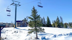 Eine Sesselbahn, ein Gebäude mit Balkon und ein mit Schnee bedeckter Hang mit Bäumen.