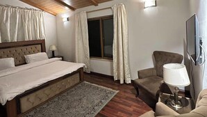 Una habitación de hotel con techo de madera, cama con cabecera, ventana con cortinas, sofá, sillón, lámpara y televisión.