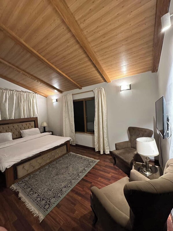 Una habitación de hotel con techo de madera, cama con cabecera, ventana con cortinas, sof á, sillón, lámpara y televisión.