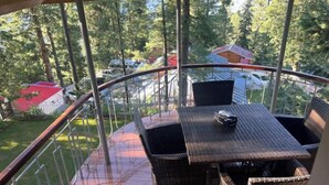 Un balcón con mobiliario de mimbre, una mesa y sillas, con vista a un área boscosa que incluye coches aparcados y un edificio con techo rojo.