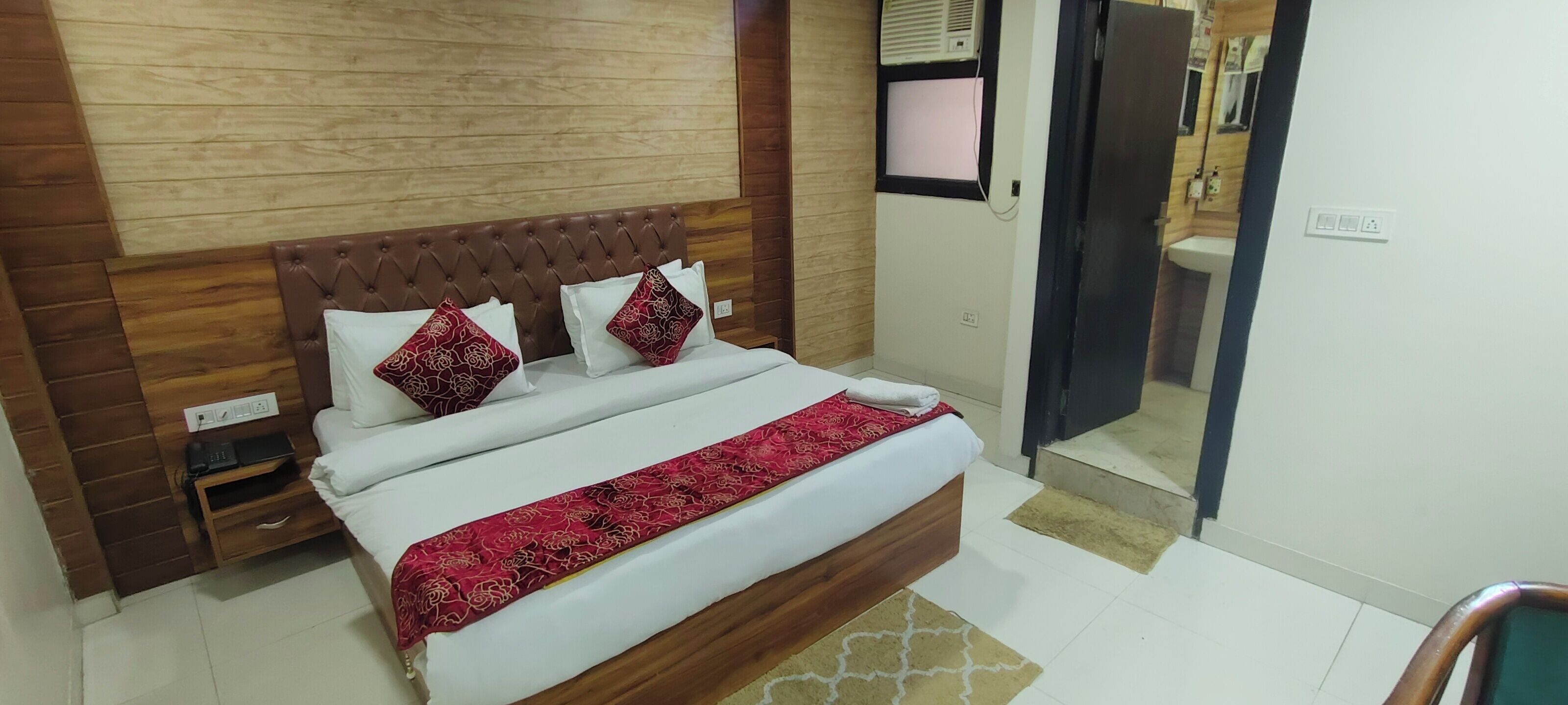 A neatly arranged bedroom with a wooden headboard, a bed with white and red bedding, a bedside table with a lamp, and a small wooden nightstand.