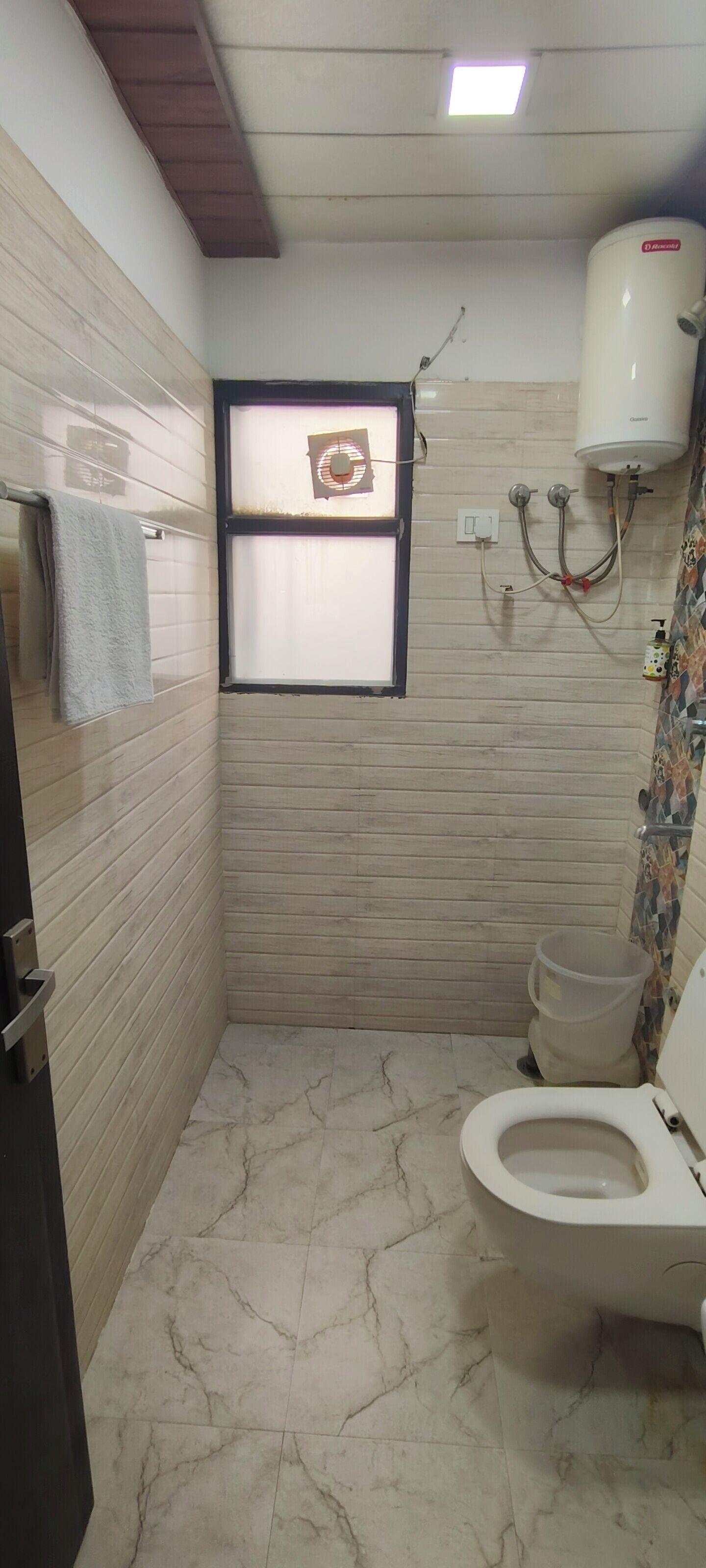 A bathroom with a toilet, a towel rack, a window with a fan, and a water heater.