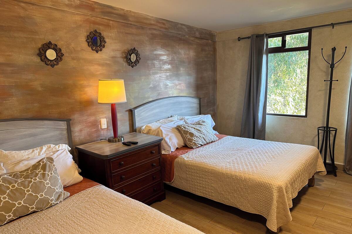 Habitación de hotel con dos camas, una mesita de noche con lámpara, perchero y ventana con cortinas.
