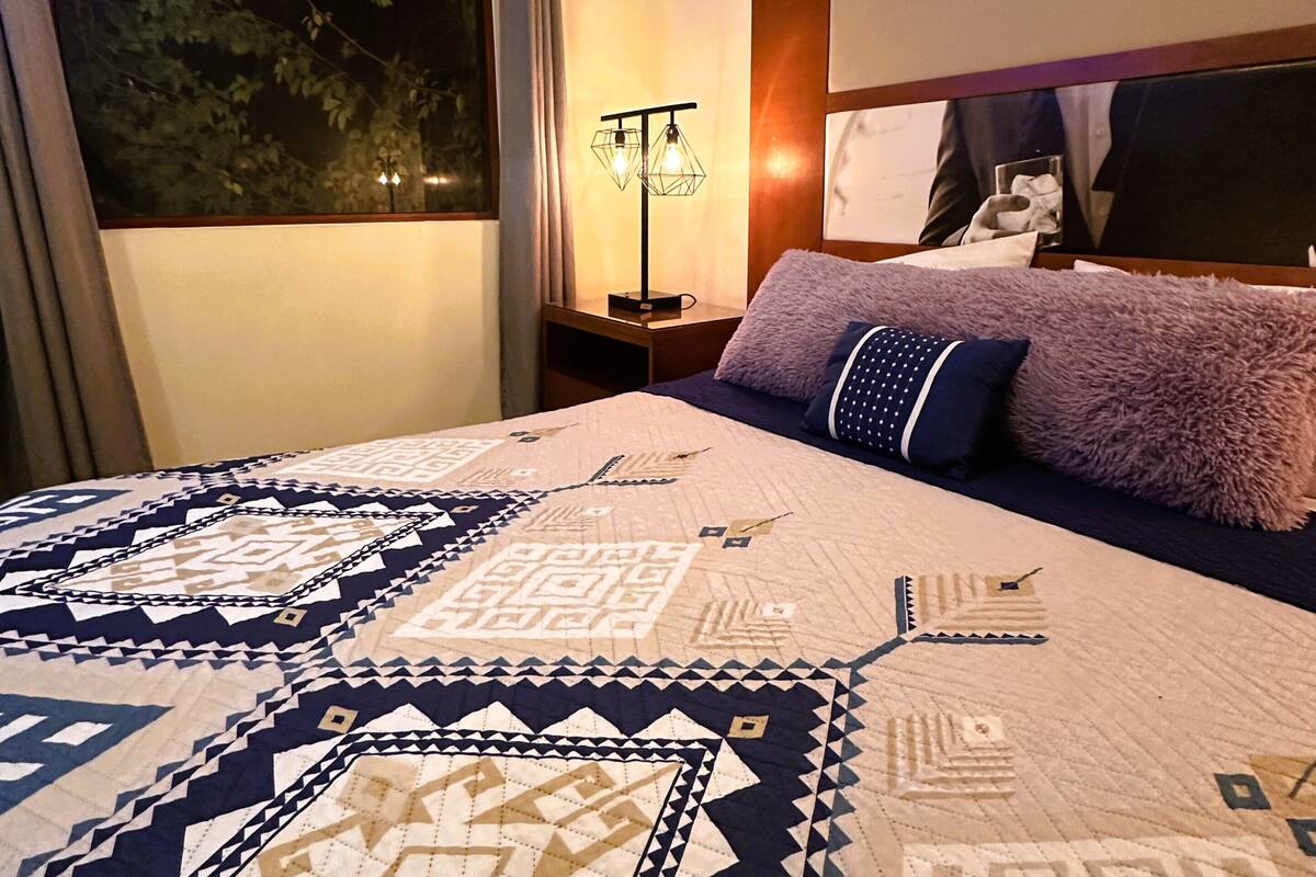 Una cama bien tendida con un edredón estampado, una lámpara de noche y una ventana con cortinas.