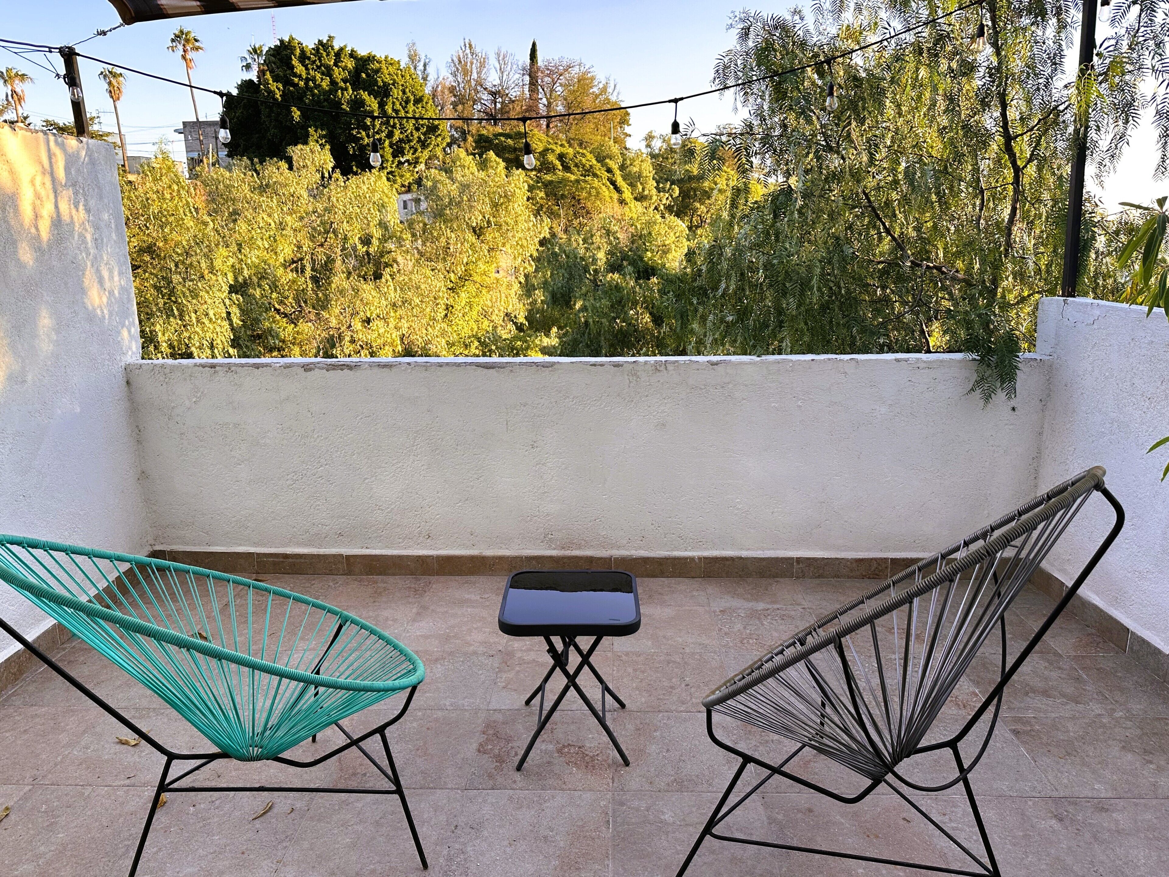 A balcony with two chairs and a small table, overlooking a garden with trees and a building in the background.