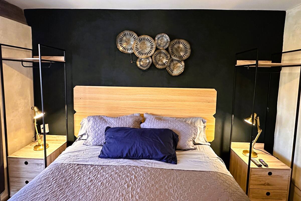 Un dormitorio moderno con cabecera de madera, cama con ropa de cama blanca y azul, dos mesitas de noche con lámparas y una decoración en la pared sobre la cama.