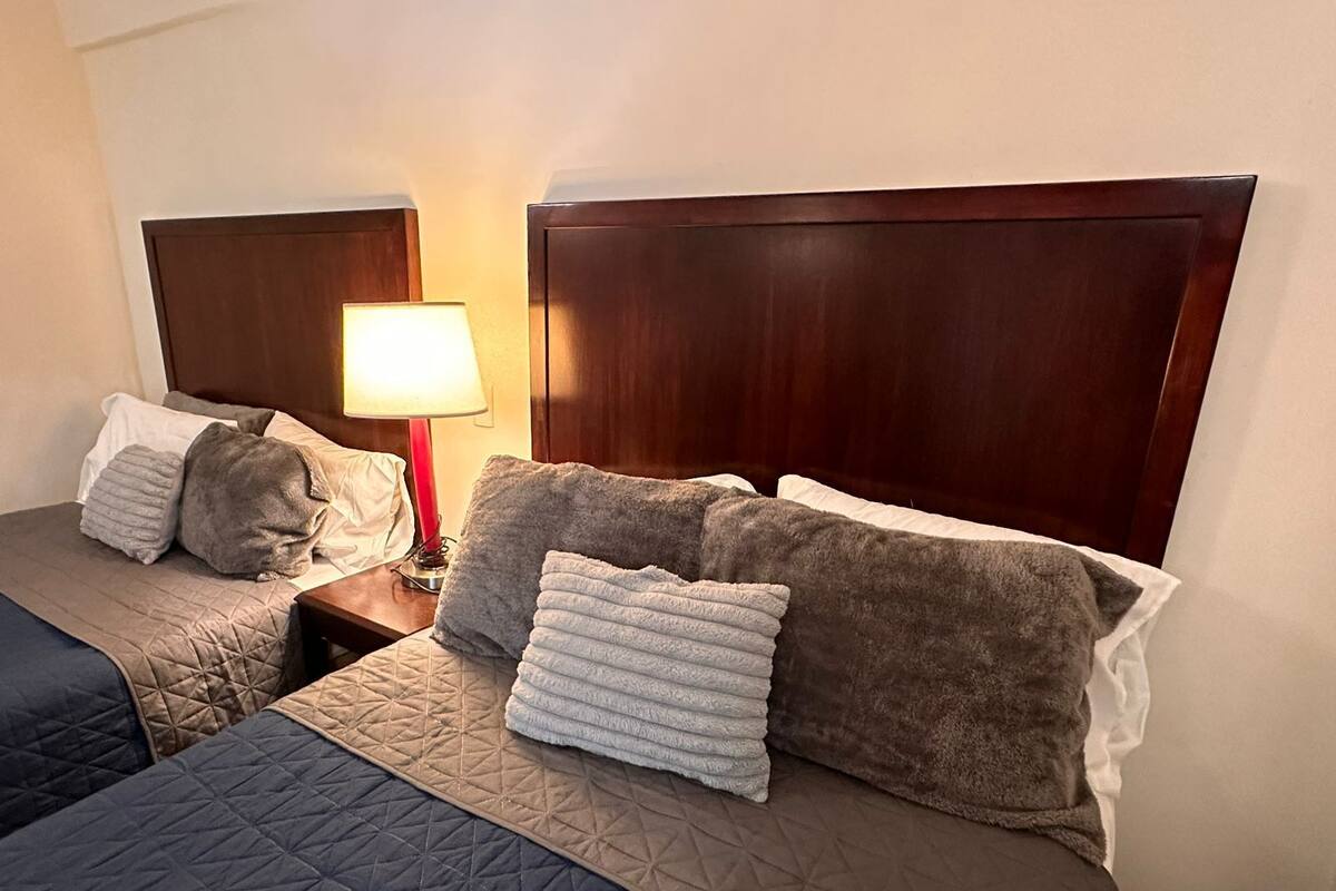 Habitación de hotel con dos camas, cada una con cabecero de madera, una mesita de noche con lámpara y varias almohadas.