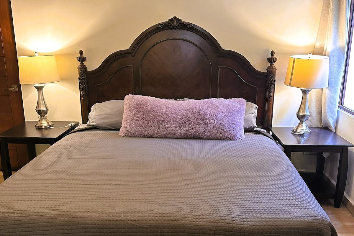 Una cama bien tendida con un cabecero, dos lámparas de noche y una alfombra estampada.