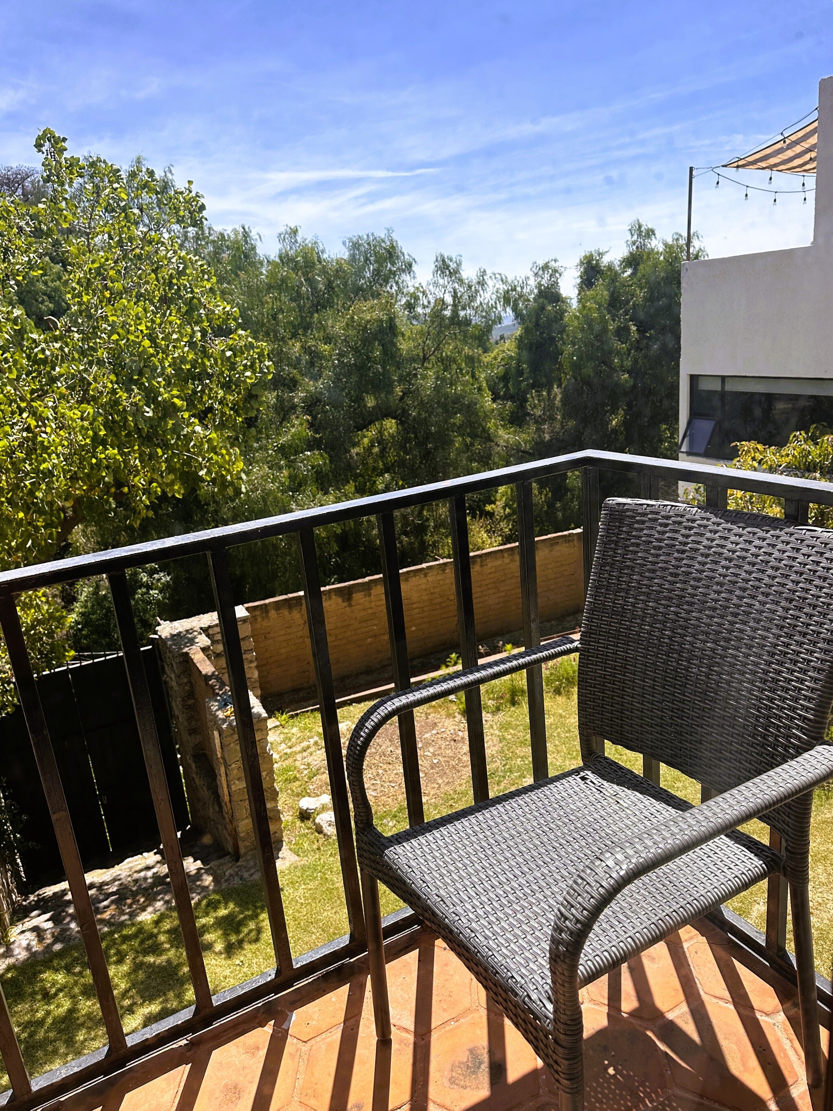 Een rieten stoel op een balkon met een metalen hekwerk, met uitzicht op een tuin en een gebouw.