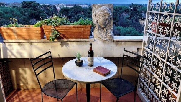 A round table with chairs, a potted plant, a wine bottle, and a book on a balcony with a view of trees and buildings. - Anno Domini Appia Antica 120 sqm, large,  elegant, close to the historic center (Roma)