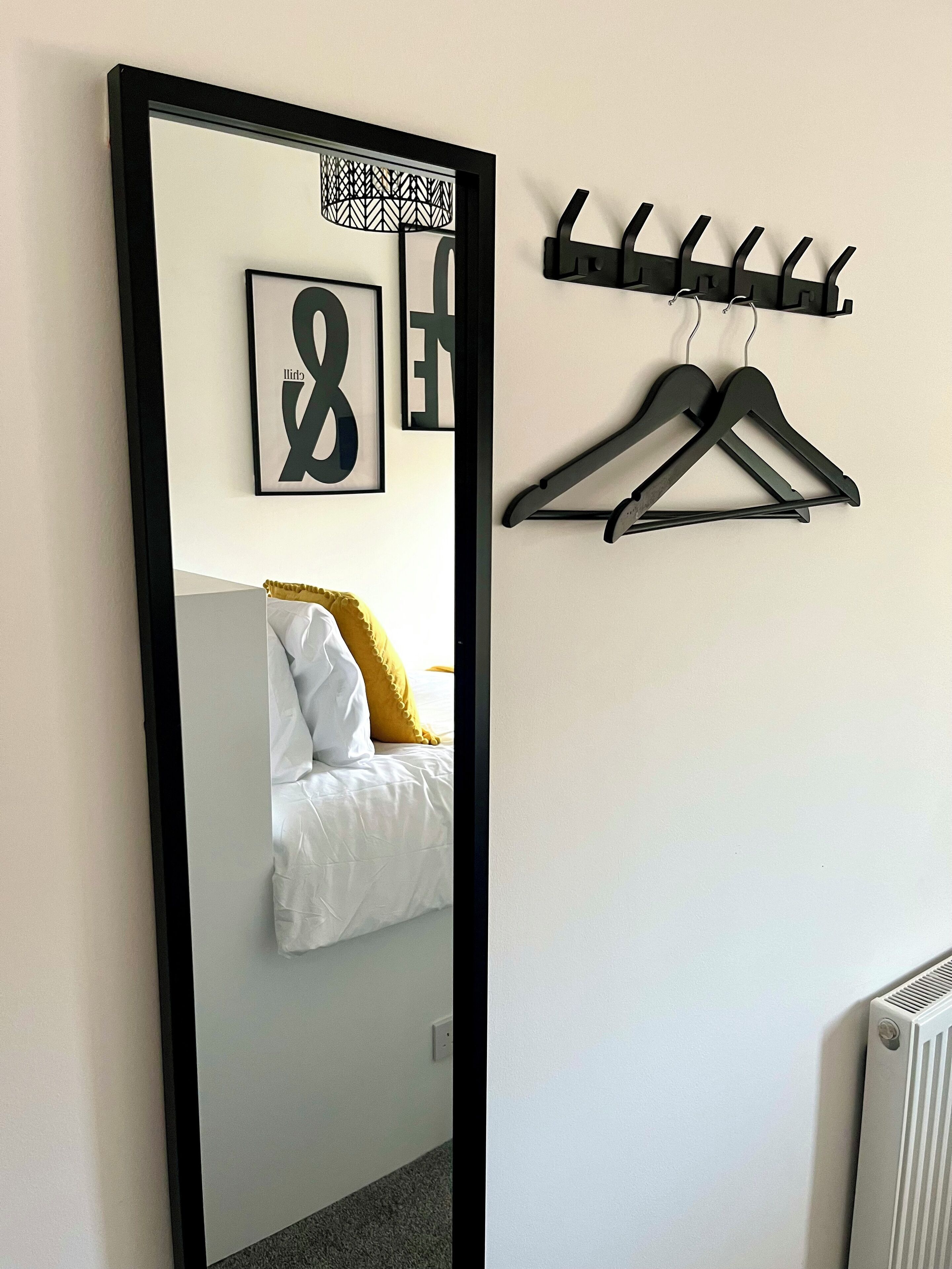 Camera da letto con un grande specchio, un letto con biancheria bianca e un cuscino giallo, un appendiabiti nero con grucce e un termosifone.