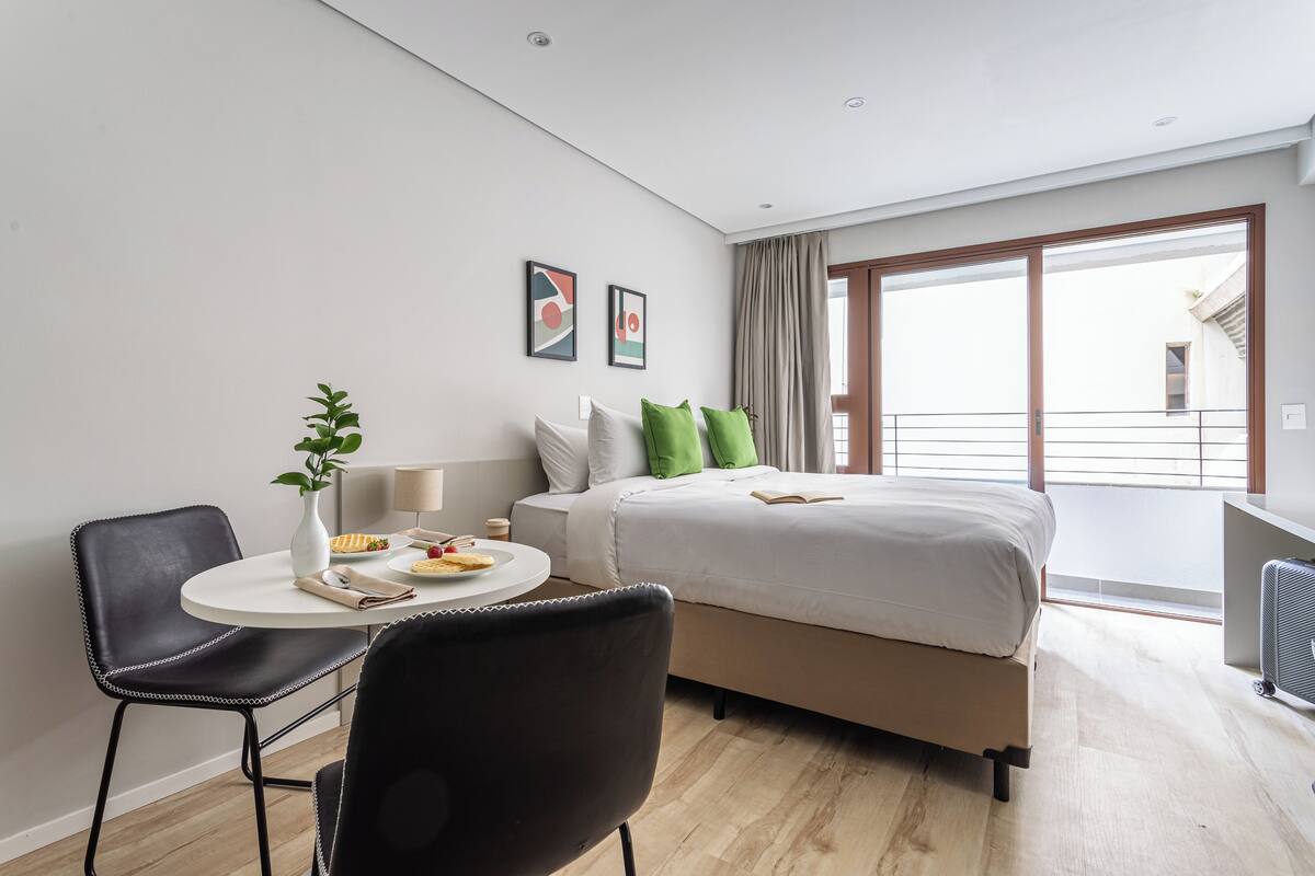 Ein modernes Hotelzimmer mit Bett, Esstisch mit Stühlen, einer kleinen Pflanze und einem Fenster mit Vorhängen.