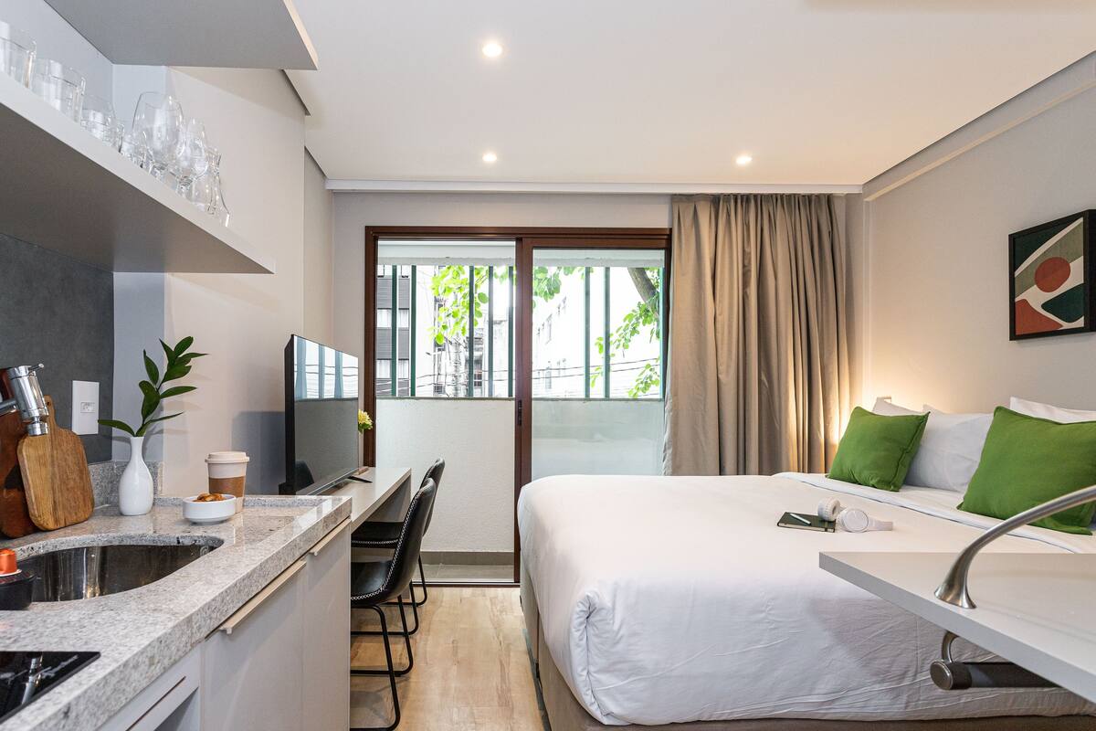 Ein modernes Hotelzimmer mit Küchenzeile, einem Bett mit grünen Kissen und Blick ins Freie.