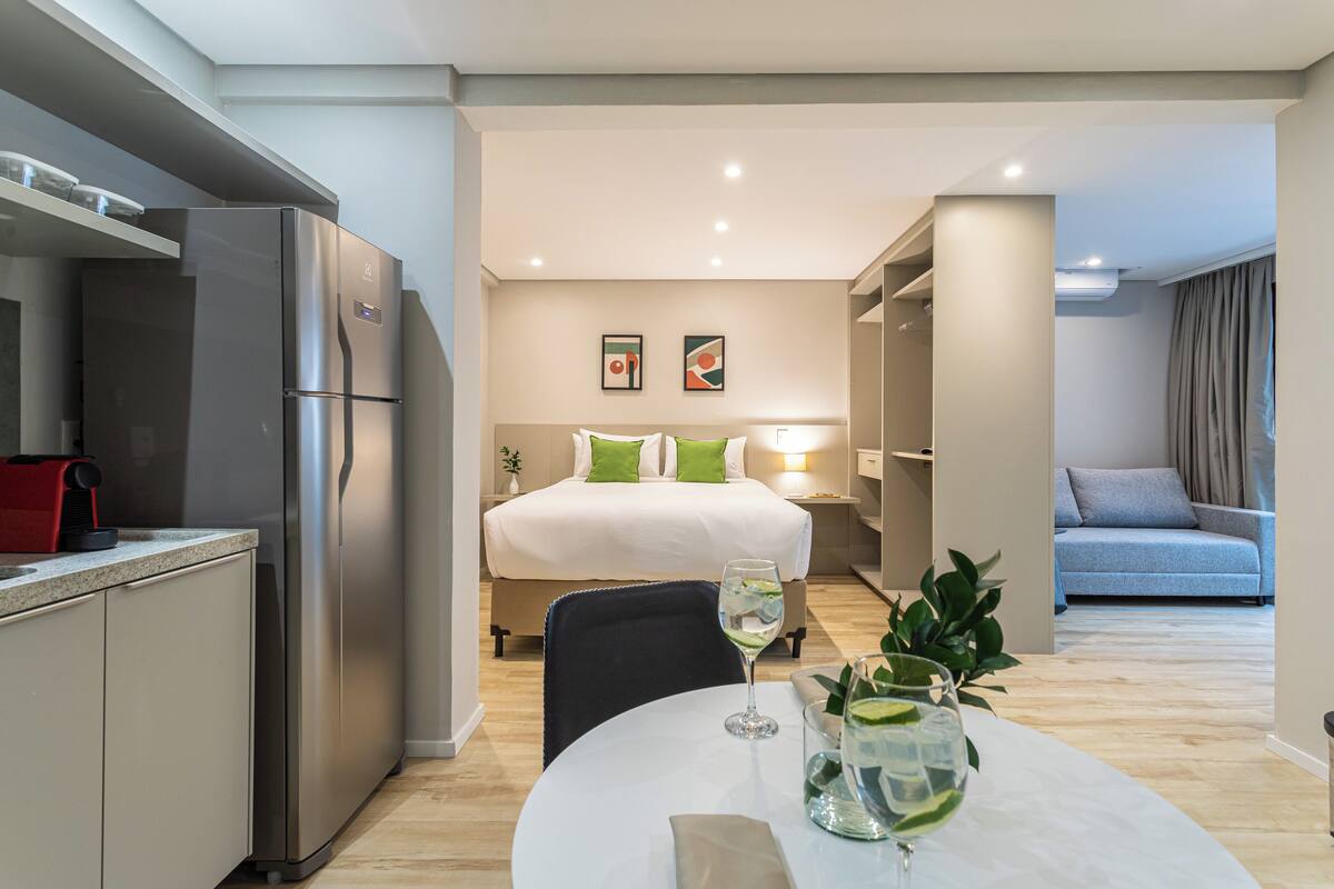 Ein modernes Hotelzimmer mit Küchenzeile, einem Esstisch und einem Glas Wasser sowie einem Bett mit grünen Kissen.