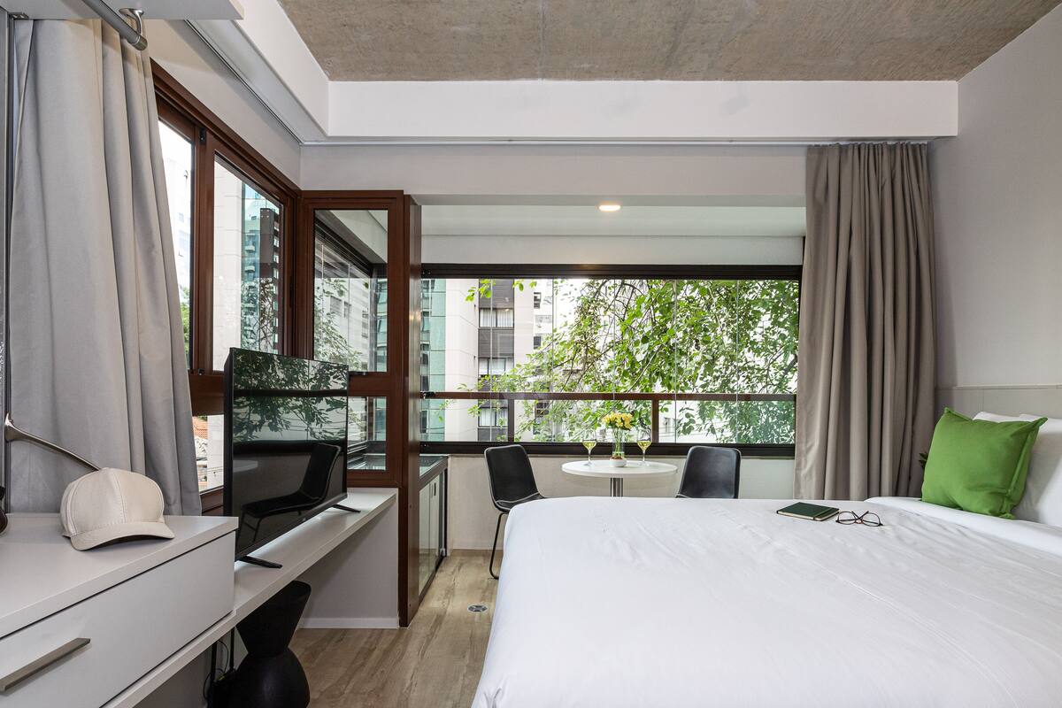 Ein Hotelzimmer mit Bett, Schreibtisch, Stuhl und einem kleinen Balkon mit Blick auf Gebäude und Bäume.
